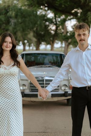 A couple in vintage-inspired attire poses together in a polka dot dress and black suit near classic cars.