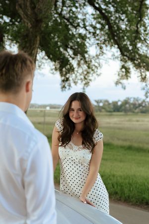 A couple in a polka dot dress and white shirt share a romantic moment under trees on a rural path.