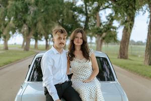 A couple poses together by a vintage car on a tree-lined country road.