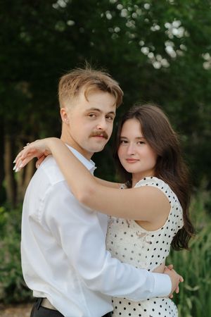 A couple embraces outdoors, with one wearing a white dress with polka dots and the other in a white shirt with a mustache.