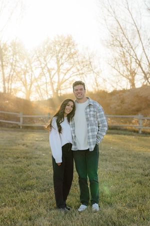 A couple stands together in a grassy field at sunset, wearing casual outfits in warm golden light.