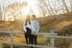 A couple stands together at sunset on a wooden fence overlooking a scenic autumn landscape.