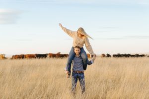 Playful piggyback ride in tall prairie grass with cattle herd visible in background.