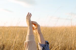 Hands raised against golden sunlit field of tall grass during sunset.