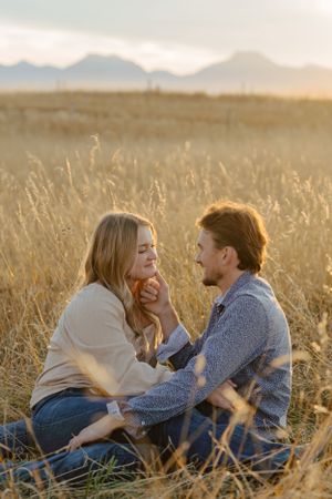 A romantic couple sitting together in tall grass with mountains in the background at golden hour.