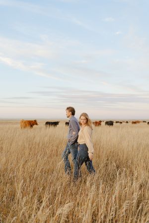 Couple standing in tall grass with grazing cattle in the background during sunset.