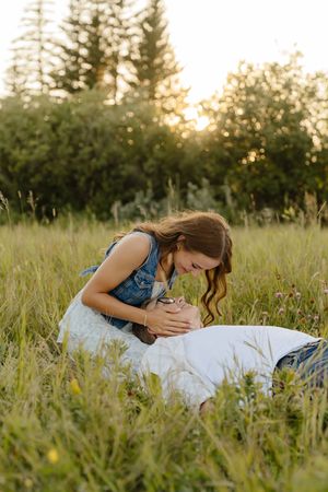 A tender moment captured in a sunlit meadow with tall grass and evergreen trees in the background at golden hour.