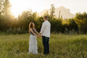 Couple holding hands walks through a sunlit meadow at golden hour with mountains in the distance.