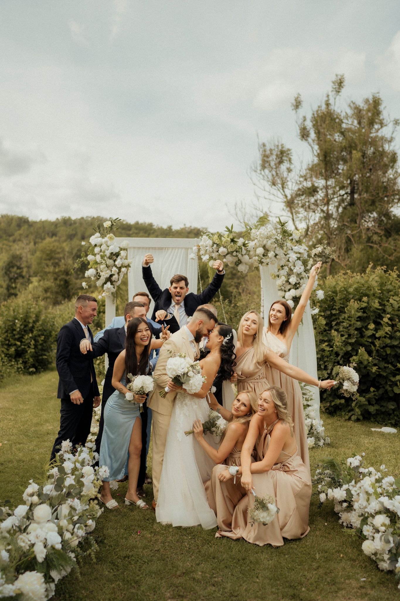 Wedding party posing enthusiastically under floral arch in outdoor garden setting.