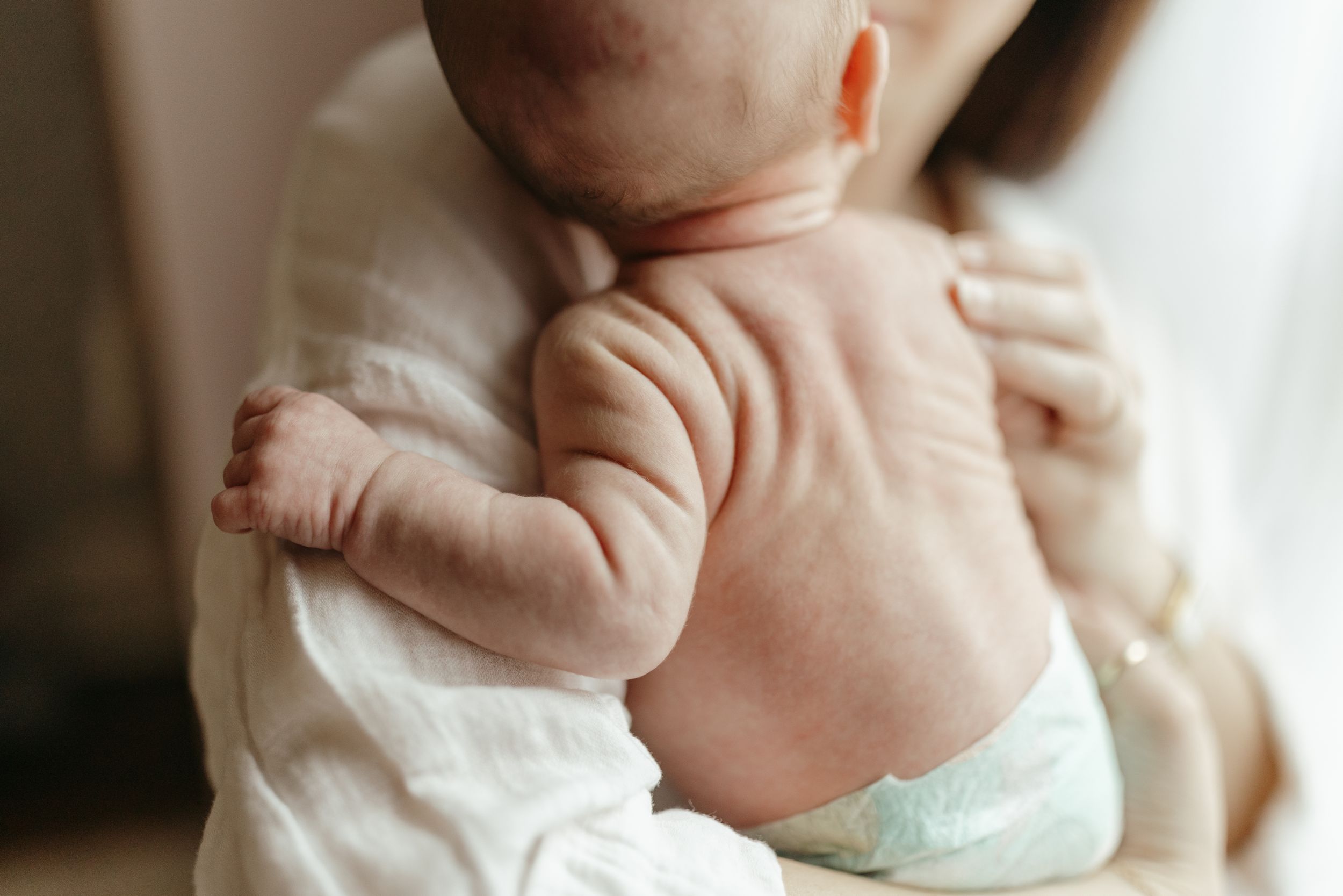 Close-up of baby's back in white onesie being held against caregiver's chest. Virginia/ North Carolina Newborn In-Home Session