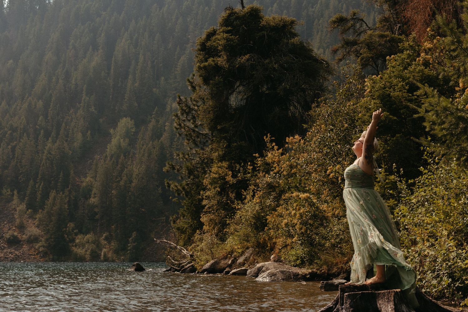 Woman in flowing green dress stands by a misty mountain lake, arms raised, surrounded by lush forest trees. Whitney Popp, AZ photographer