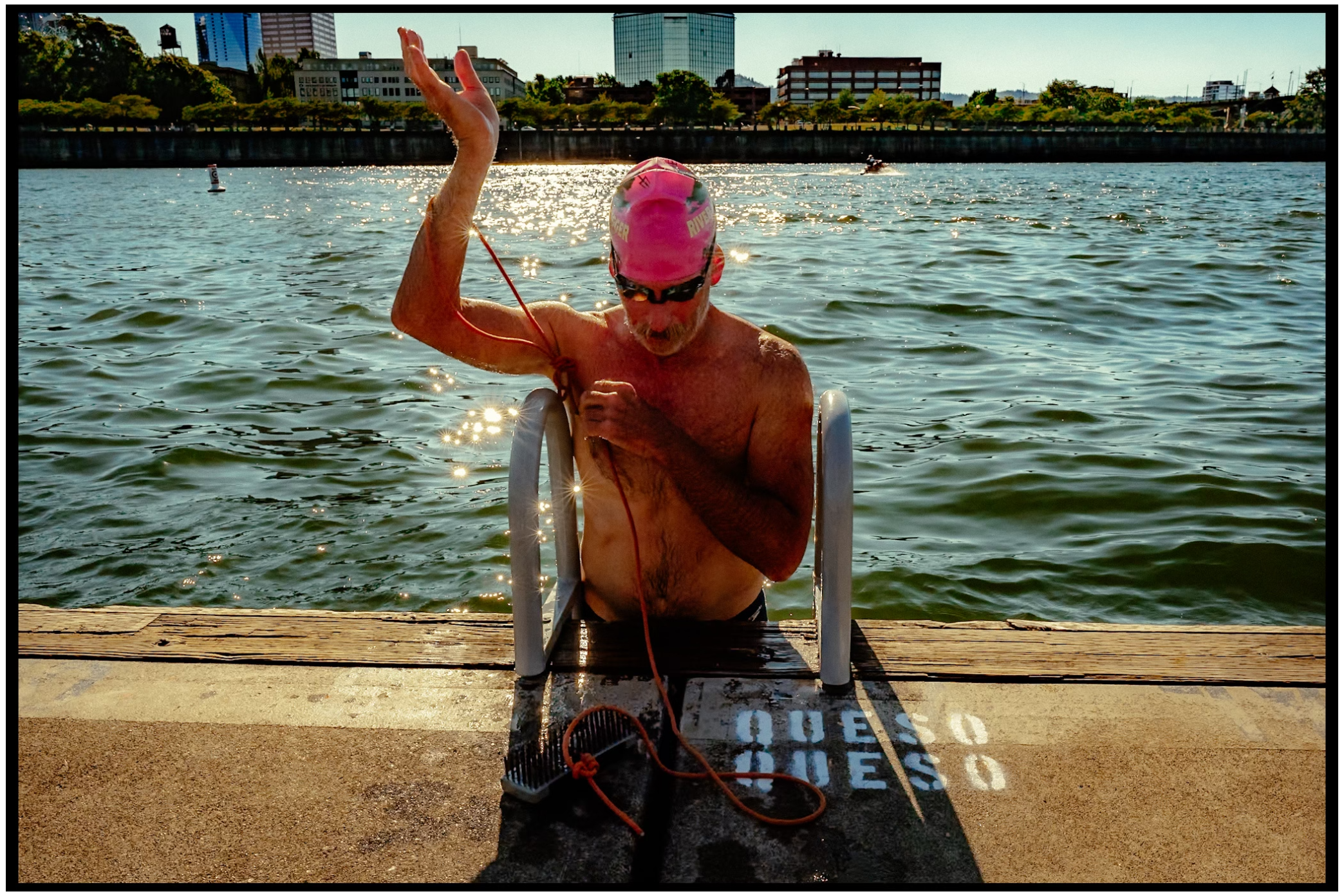 A figure in a pink hat on a dock ladder by a waterfront, raising their arm. Willamette River, Portland, OR