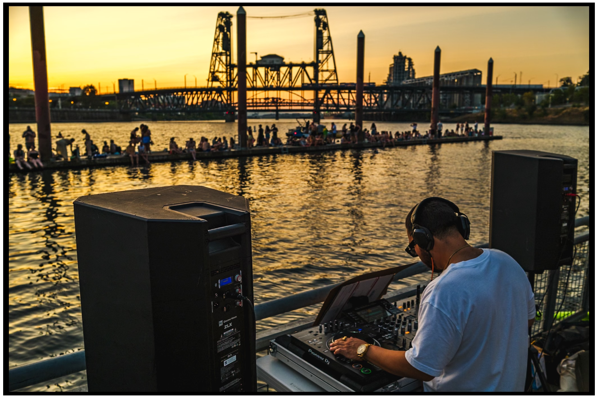 DJ performs at outdoor waterfront event with sunset bridge silhouette in background. Willamette River, Portland, OR