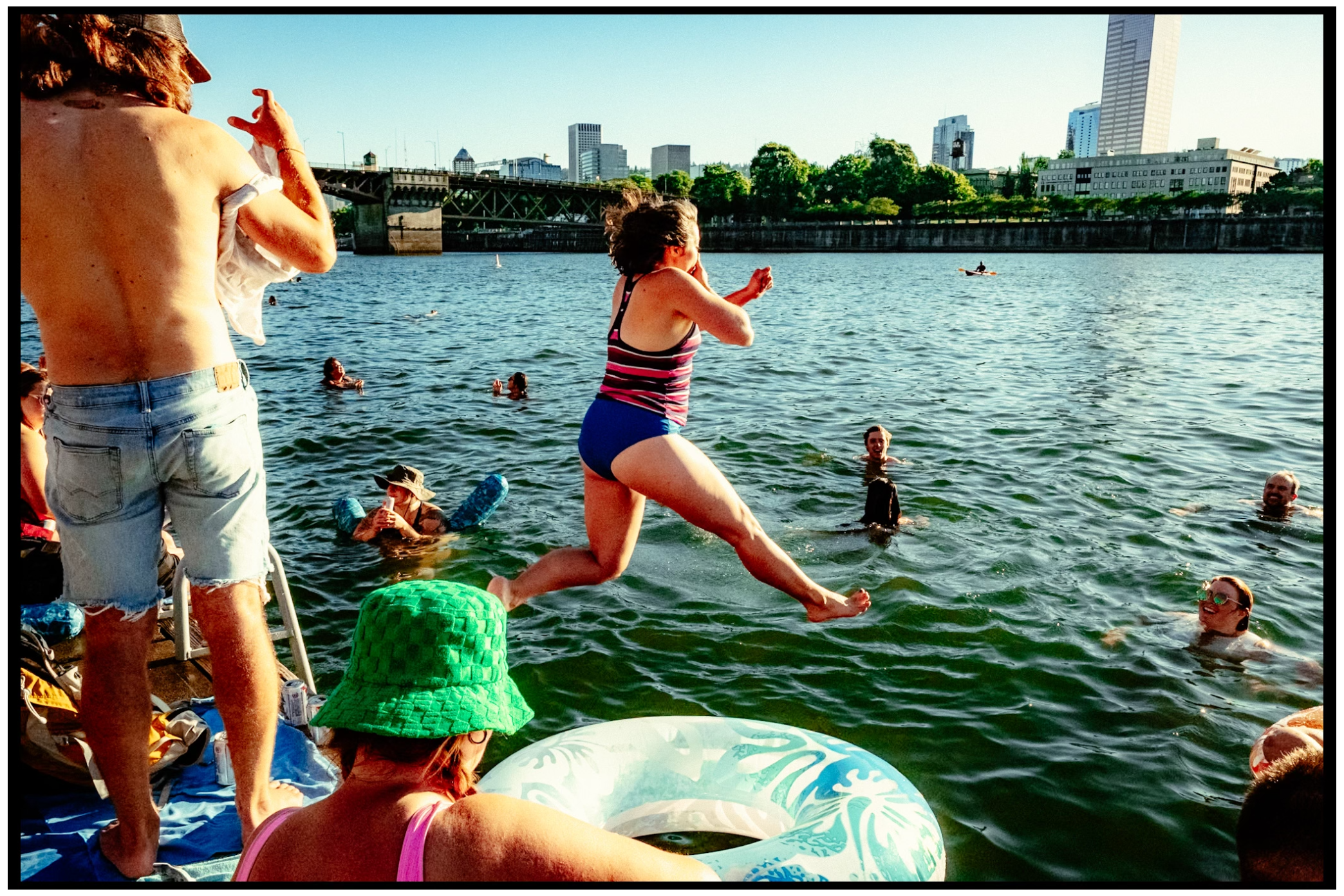Person jumping into a river while others swim during a summer day in the city. Willamette River, Portland, OR