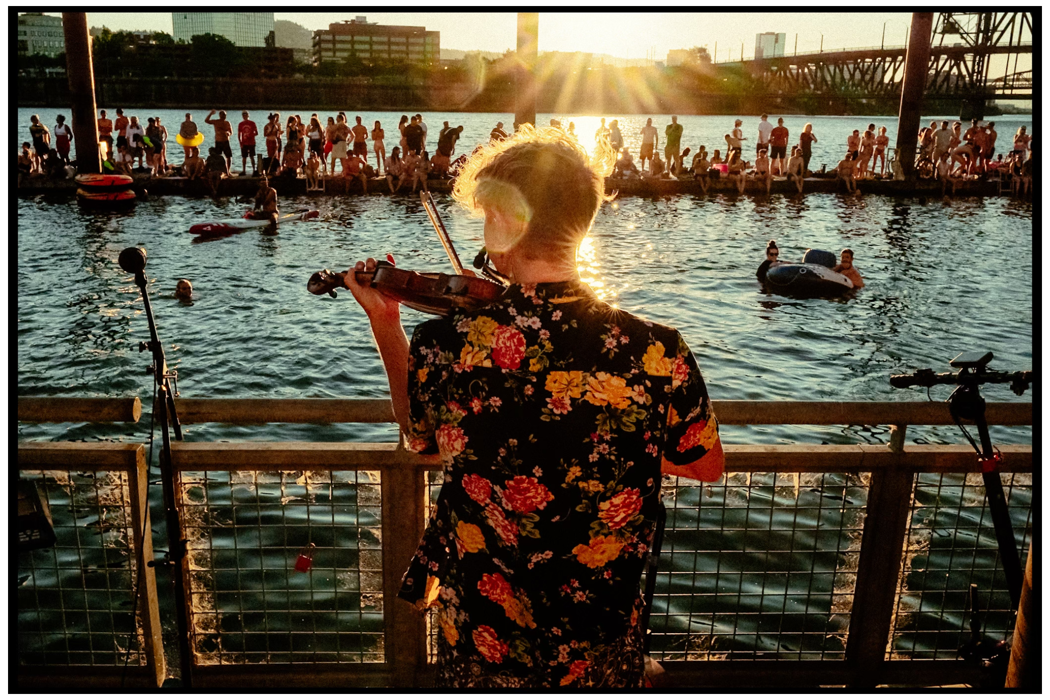 Person in floral shirt watches sunset over crowded waterfront gathering near industrial bridge structure. Willamette River, Portland, OR