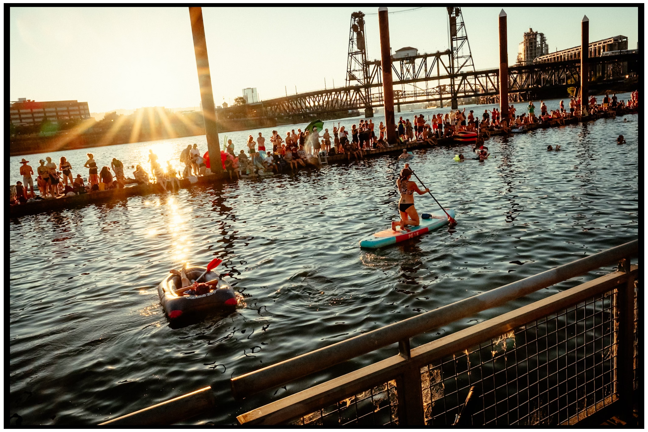 People swimming and paddleboarding at sunset beneath industrial bridge structure. Willamette River, Portland, OR