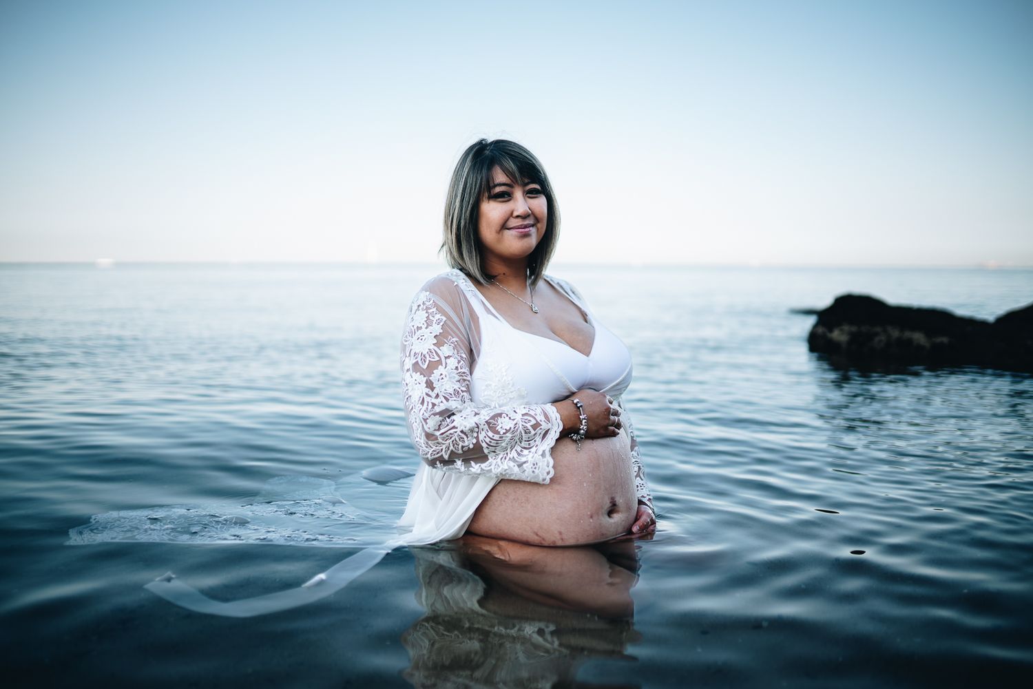 Person in white lace dress poses for maternity photo while standing in calm ocean water taken by a Melbourne maternity photographer