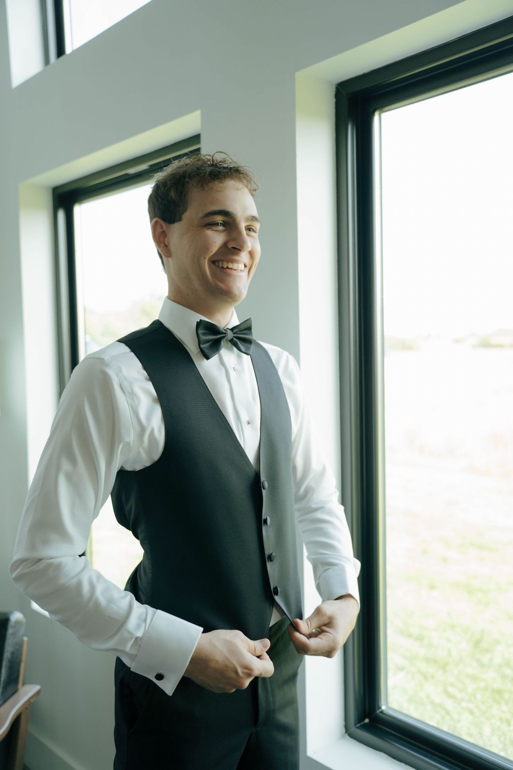 A groom in formal attire with black vest and bowtie stands by a window smiling.