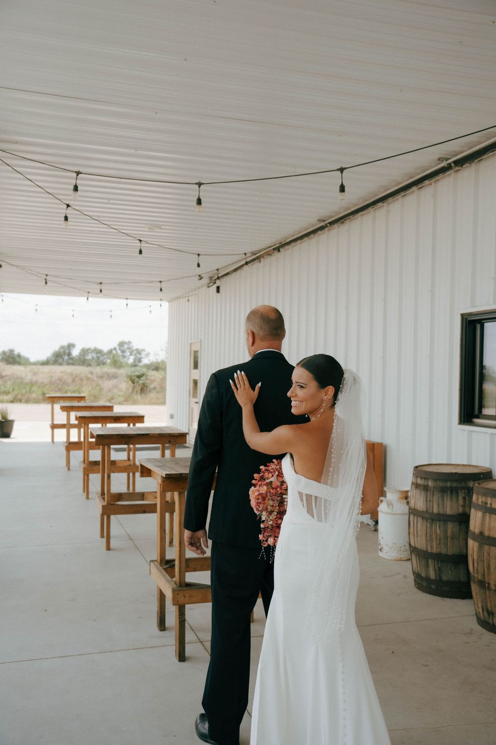 Couple shares intimate moment against white barn wall with string lights and rustic barrels.
