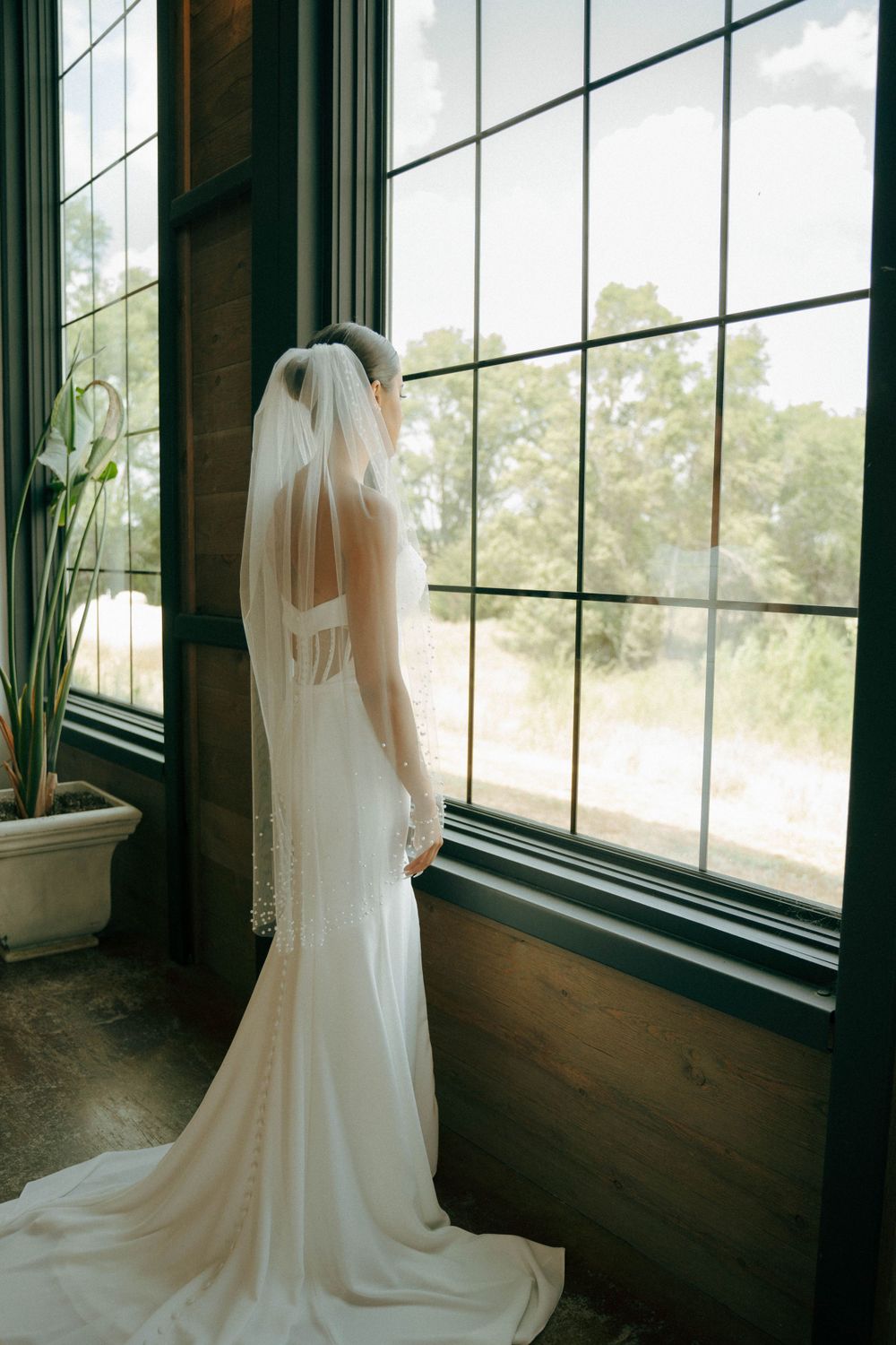 Bride in elegant white gown and veil stands silhouetted against large window in romantic moment.