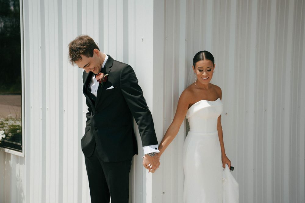 A couple holds hands while standing on opposite sides of a white wall during their wedding first look.