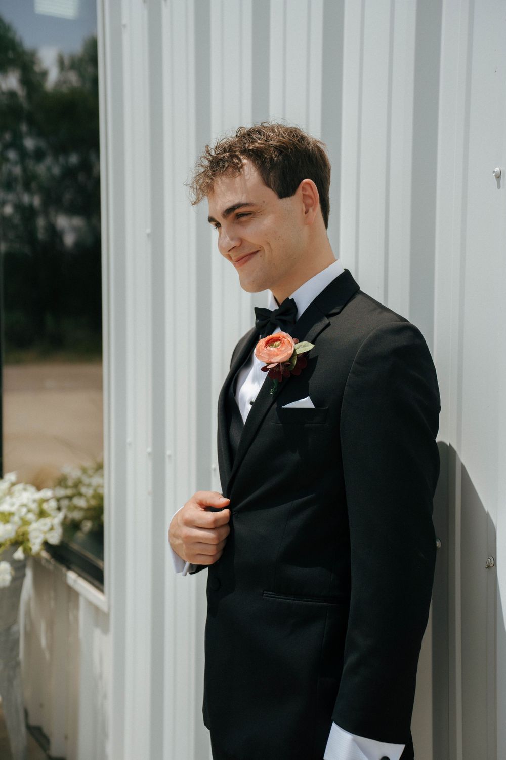 A groom in a black tuxedo with coral boutonniere smiles while standing against a white wall.
