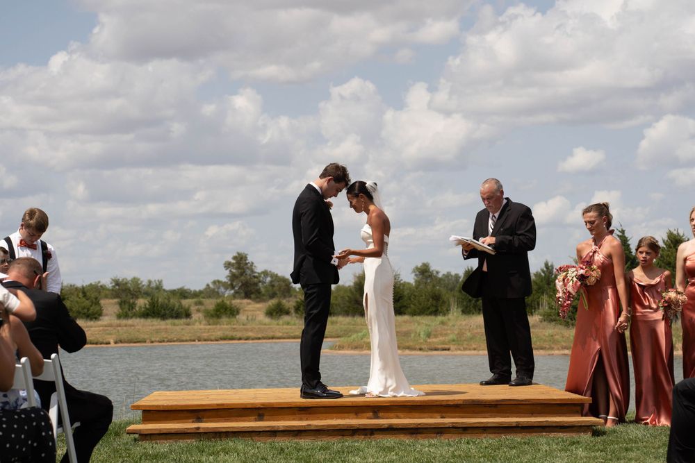 A couple exchanges vows on a wooden platform by a lake during their outdoor wedding ceremony.