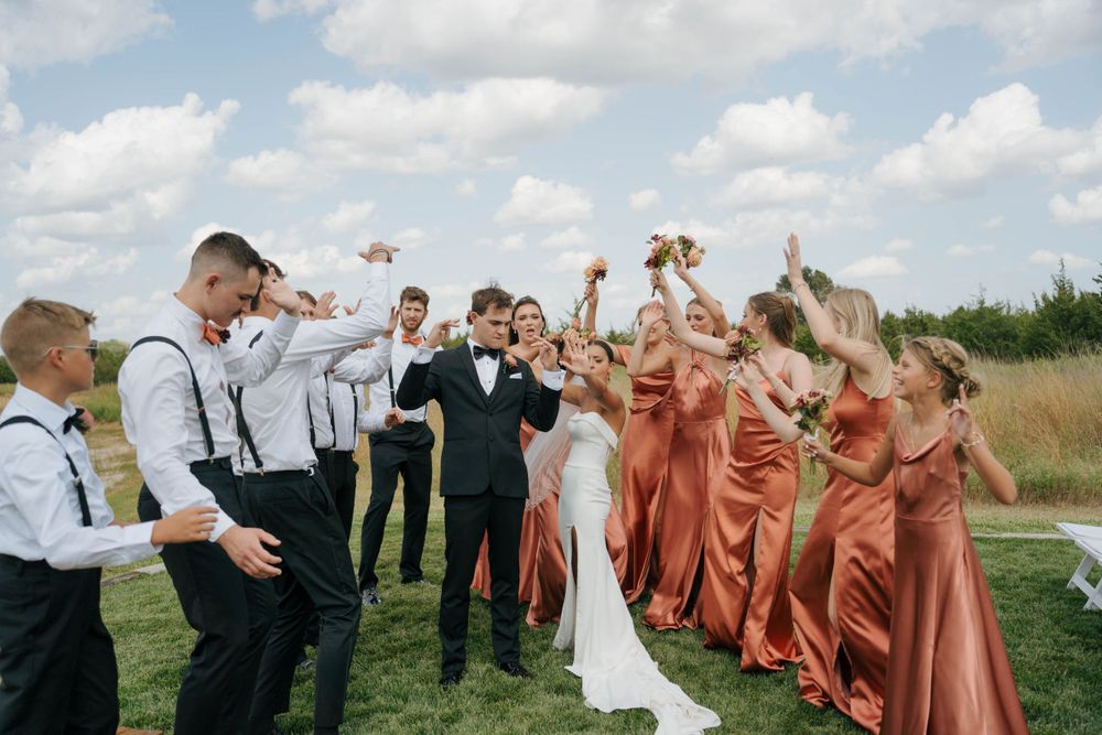 Wedding party members cheer and celebrate outdoors in coral-colored attire.