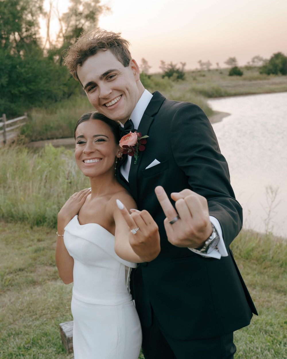 A playful wedding couple making hand gestures and smiling outdoors by a waterfront.