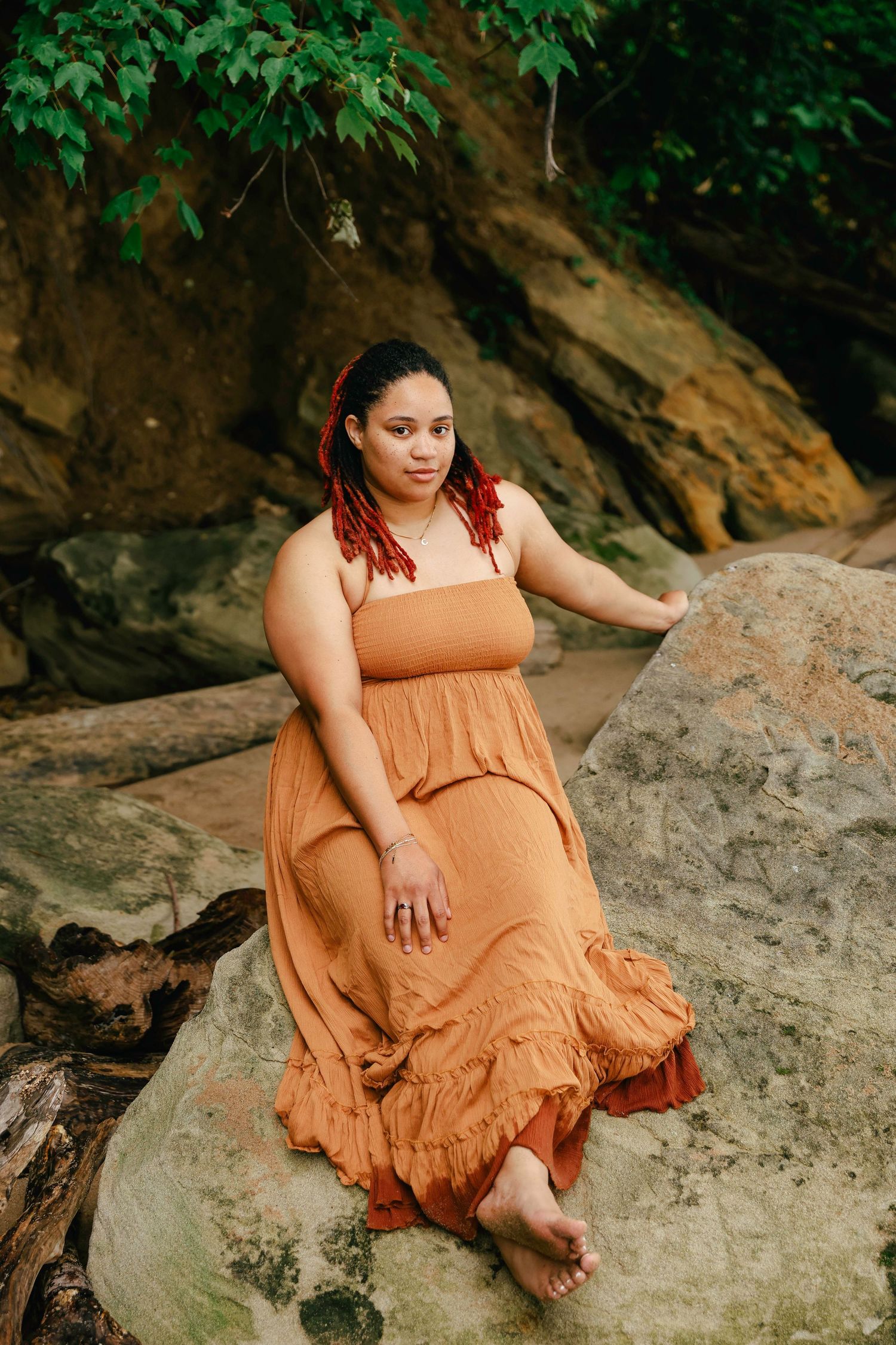 A person in a flowing orange dress sits on a large rock formation surrounded by green foliage.