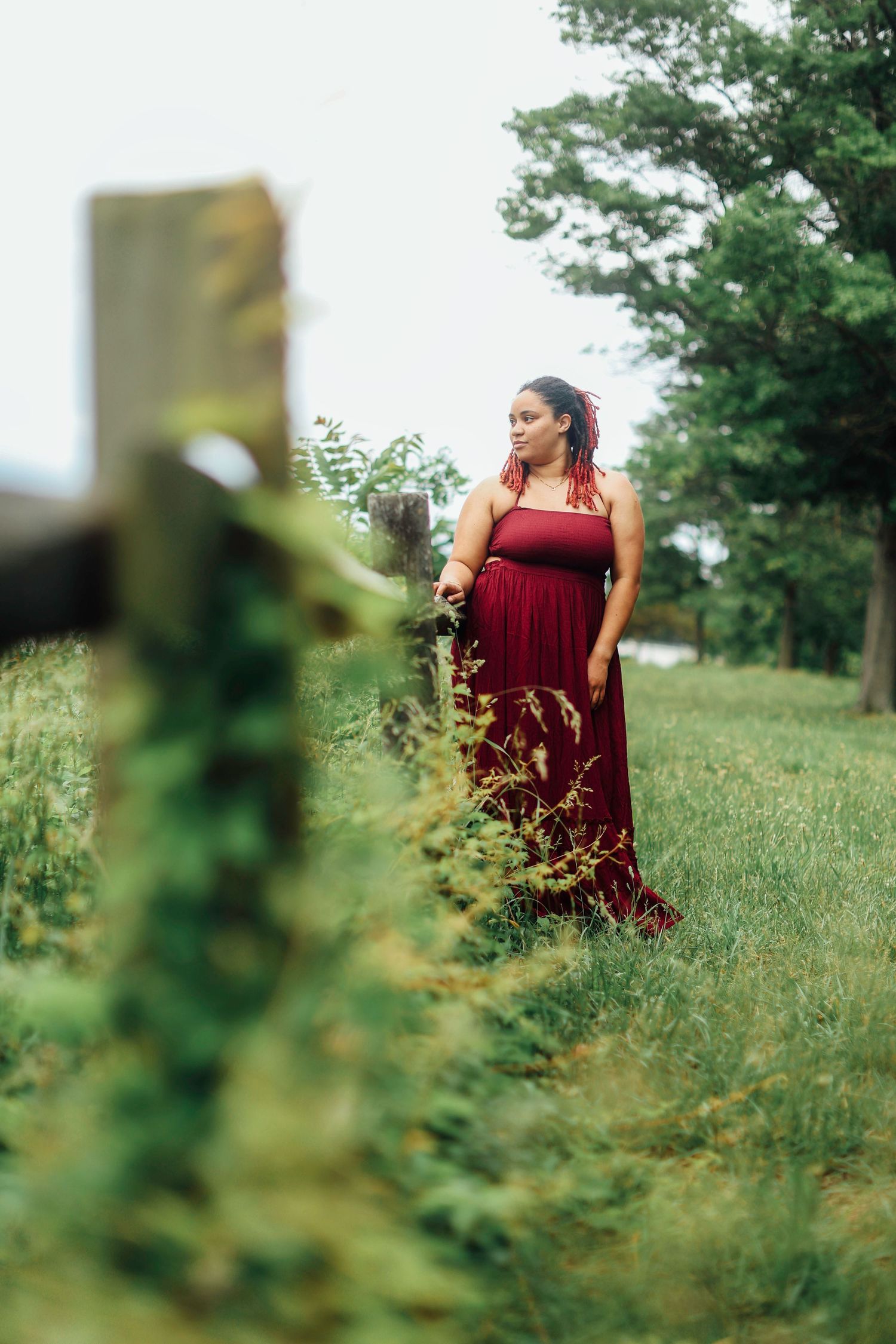 A person in a flowing burgundy dress stands among green foliage in a sunlit meadow, photographed from different angles.