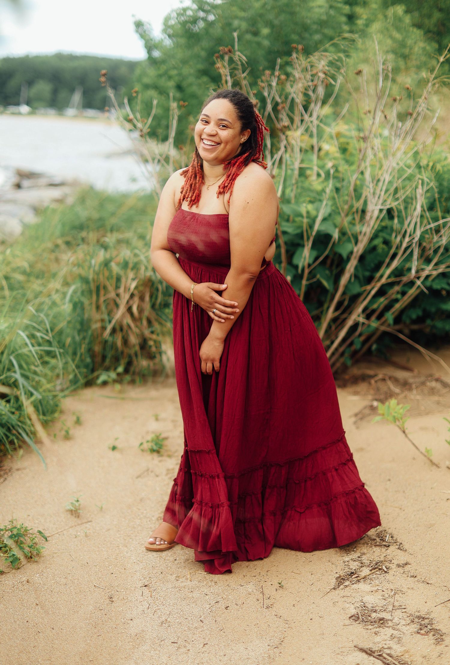 Woman in flowing burgundy maxi dress laughs joyfully while posing on sandy beach near lake and tall grass.
