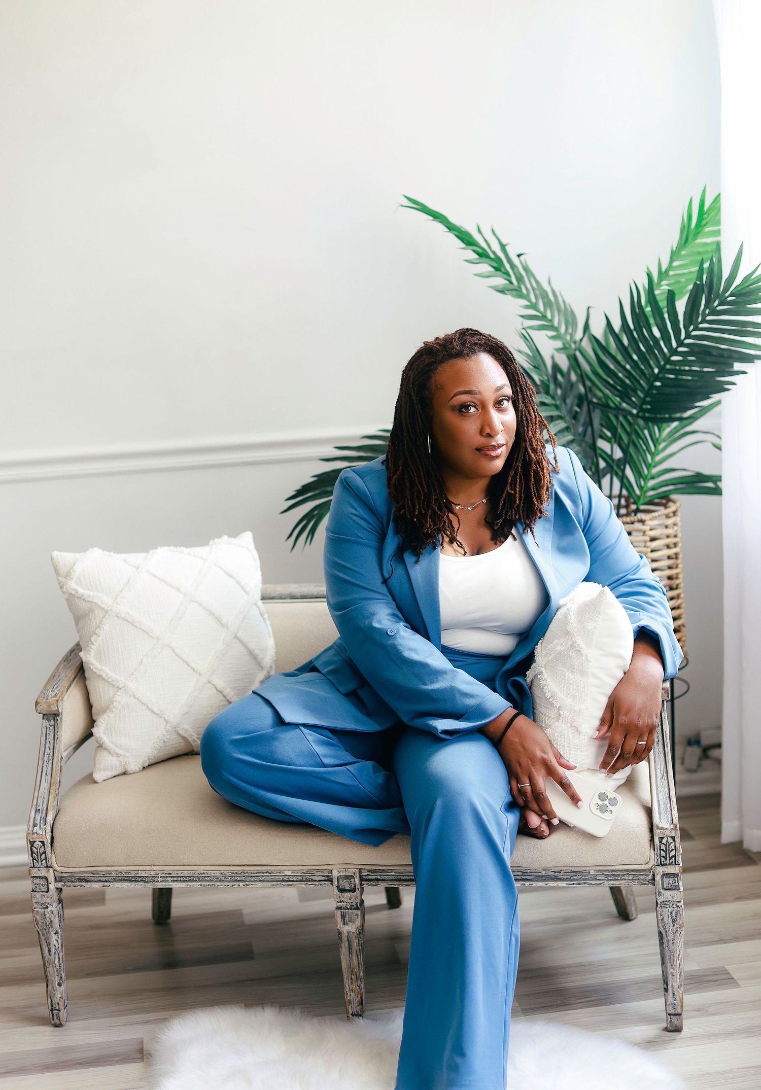 Person in a blue blazer and white top lounges gracefully on a vintage-style settee with white cushions and palm decor.