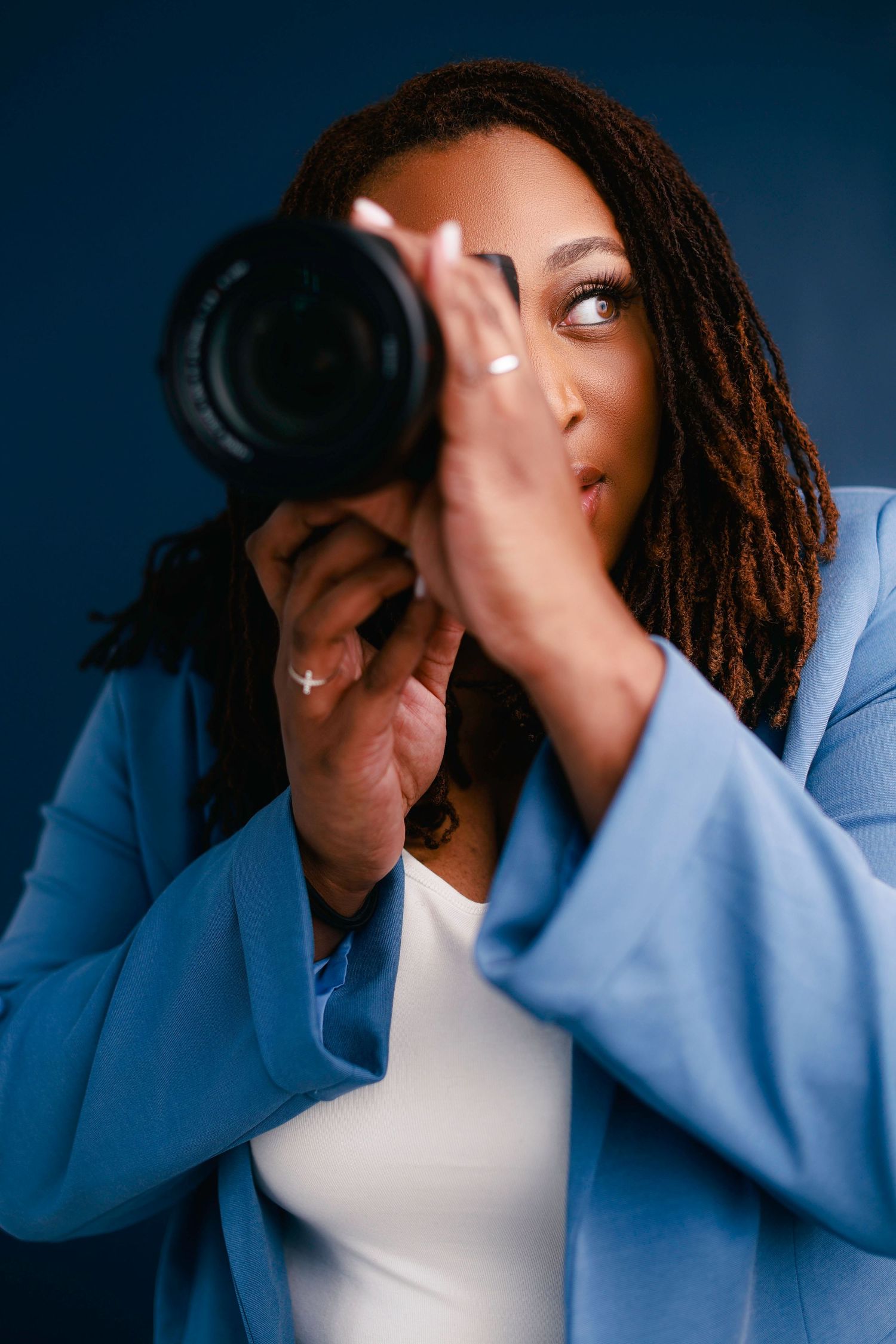 Professional photographer wearing blue blazer and white top focuses intently while shooting with DSLR camera against dark backdrop.