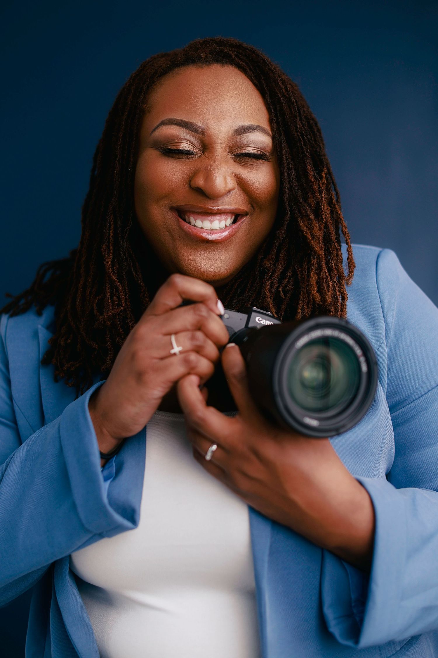 Professional photographer in a light blue blazer smiles joyfully while holding a DSLR camera against blue backdrop.
