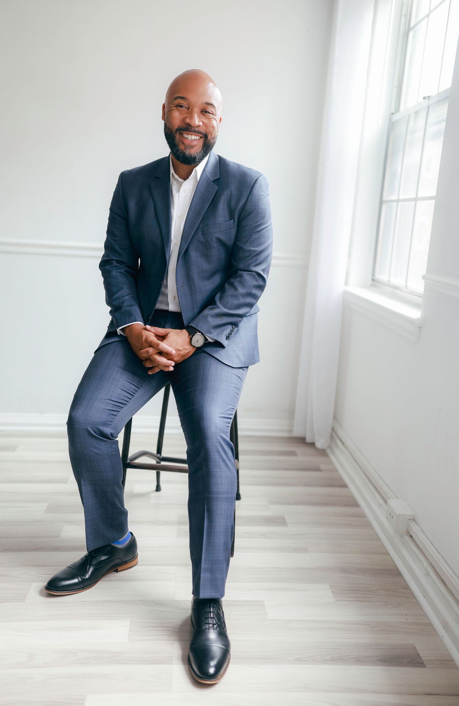 Professional portrait in a bright room showing someone seated on a stool wearing a navy suit with white shirt and dress shoes.