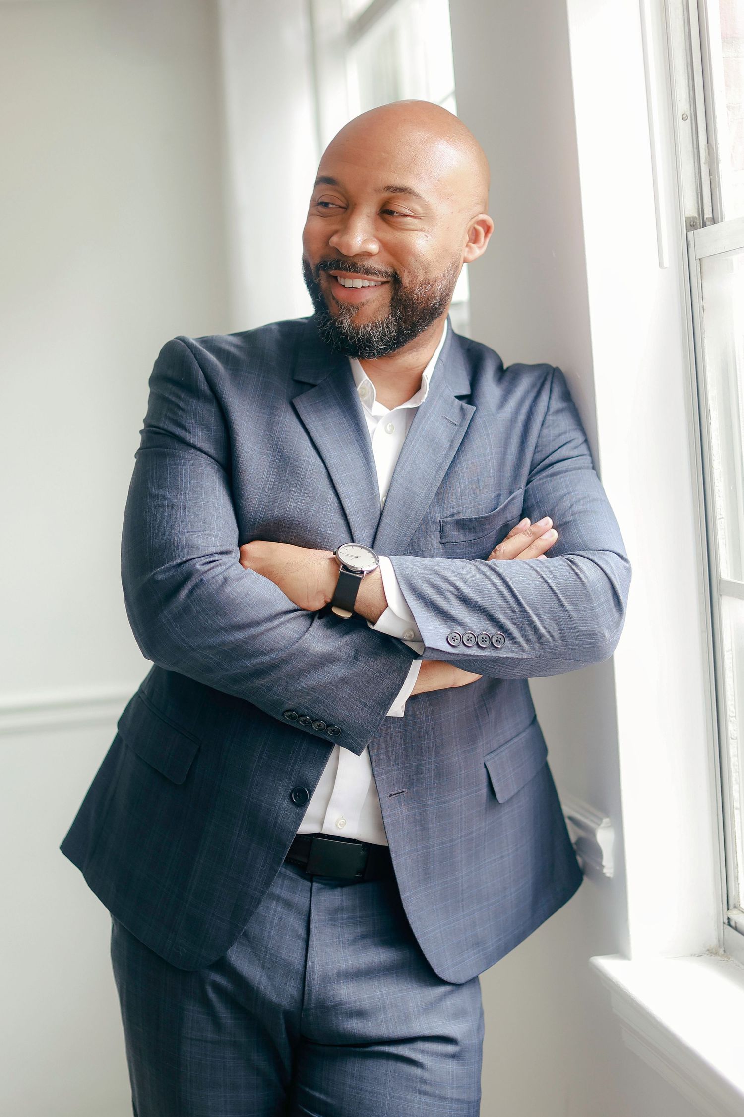 Professional in a well-fitted grey suit stands by window with arms crossed and a warm smile.