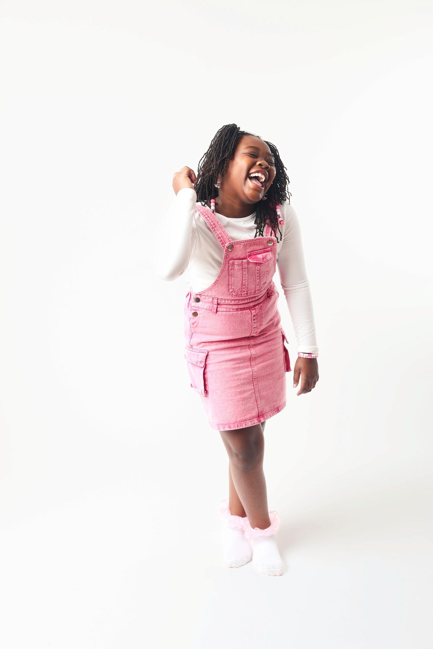A child wearing a pink overall dress and white shirt laughs and plays against a white studio backdrop.