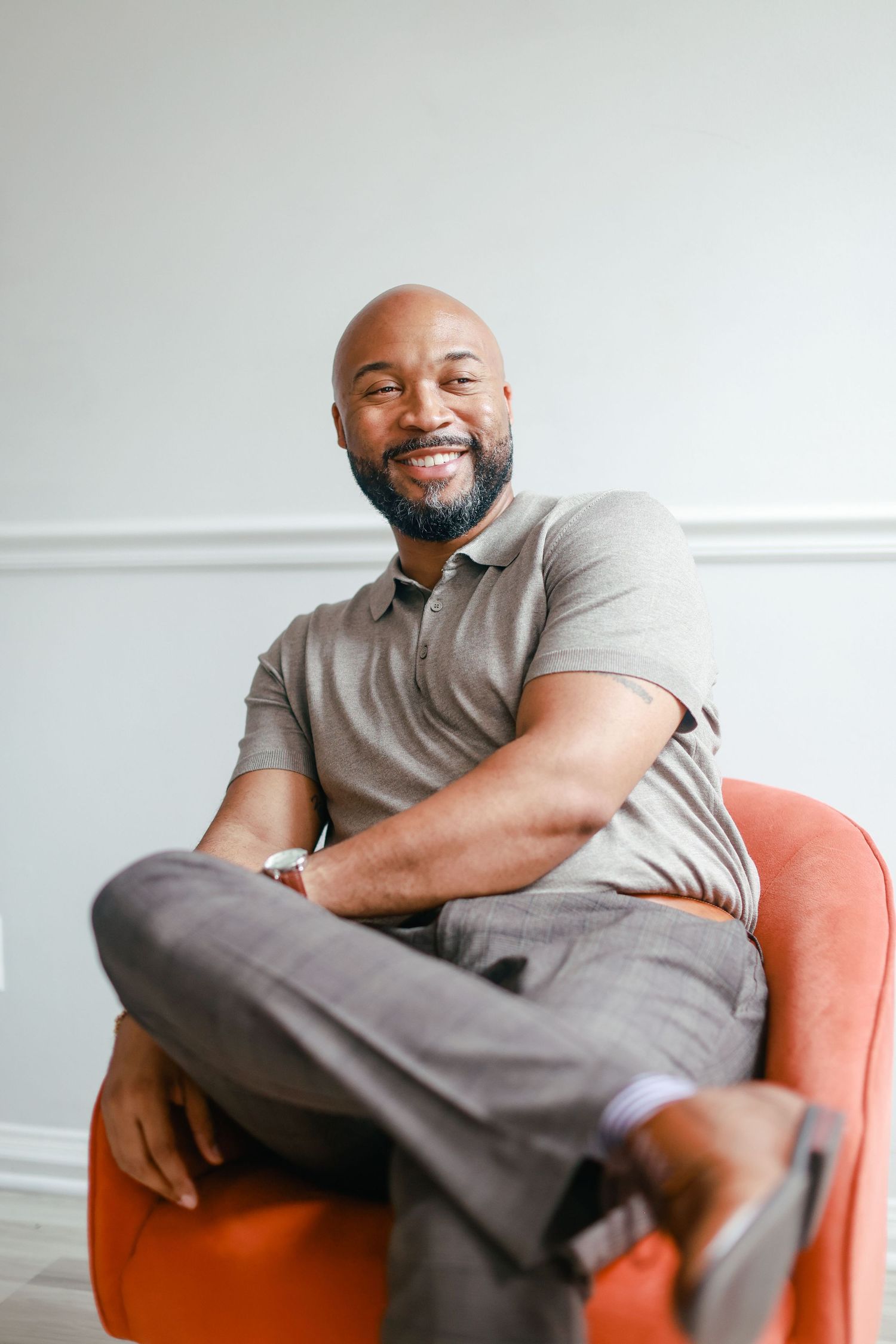 Person in grey attire sits relaxed and smiling in an orange chair against a white wall.