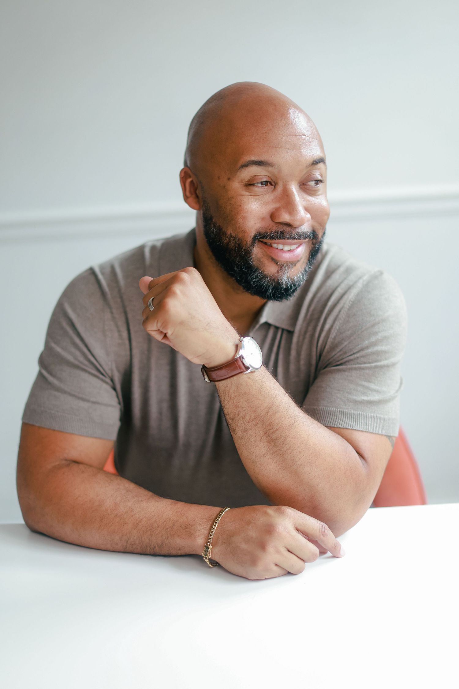 A smiling person in a grey t-shirt leans on a white table while wearing a wristwatch.