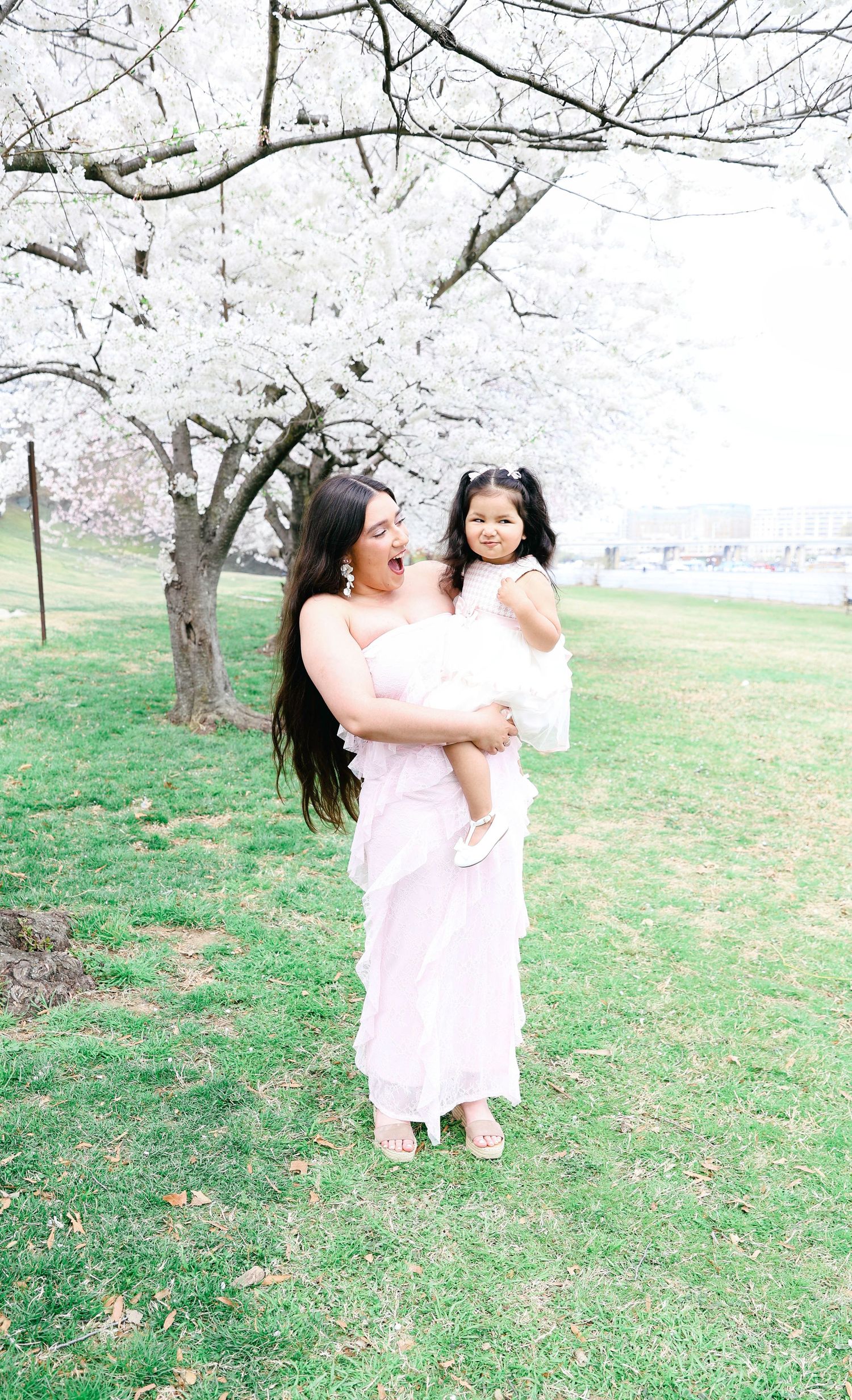 Two people in flowing white dresses share a joyful moment under blooming cherry blossom trees in a spring garden.