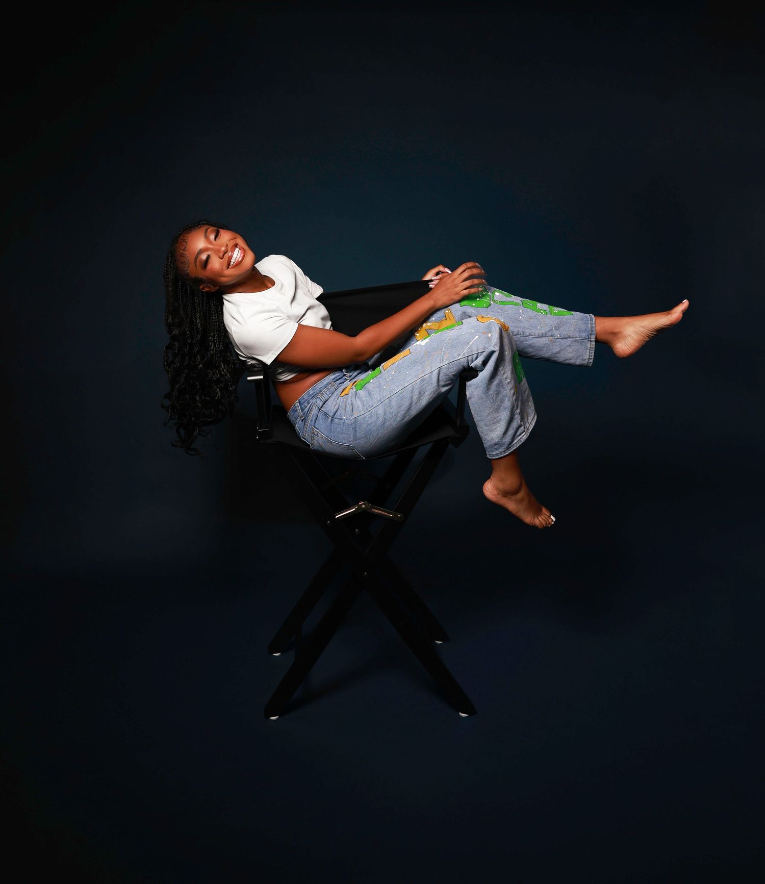 Joyful person in white t-shirt and jeans reclines playfully on a black chair against a dark studio background.