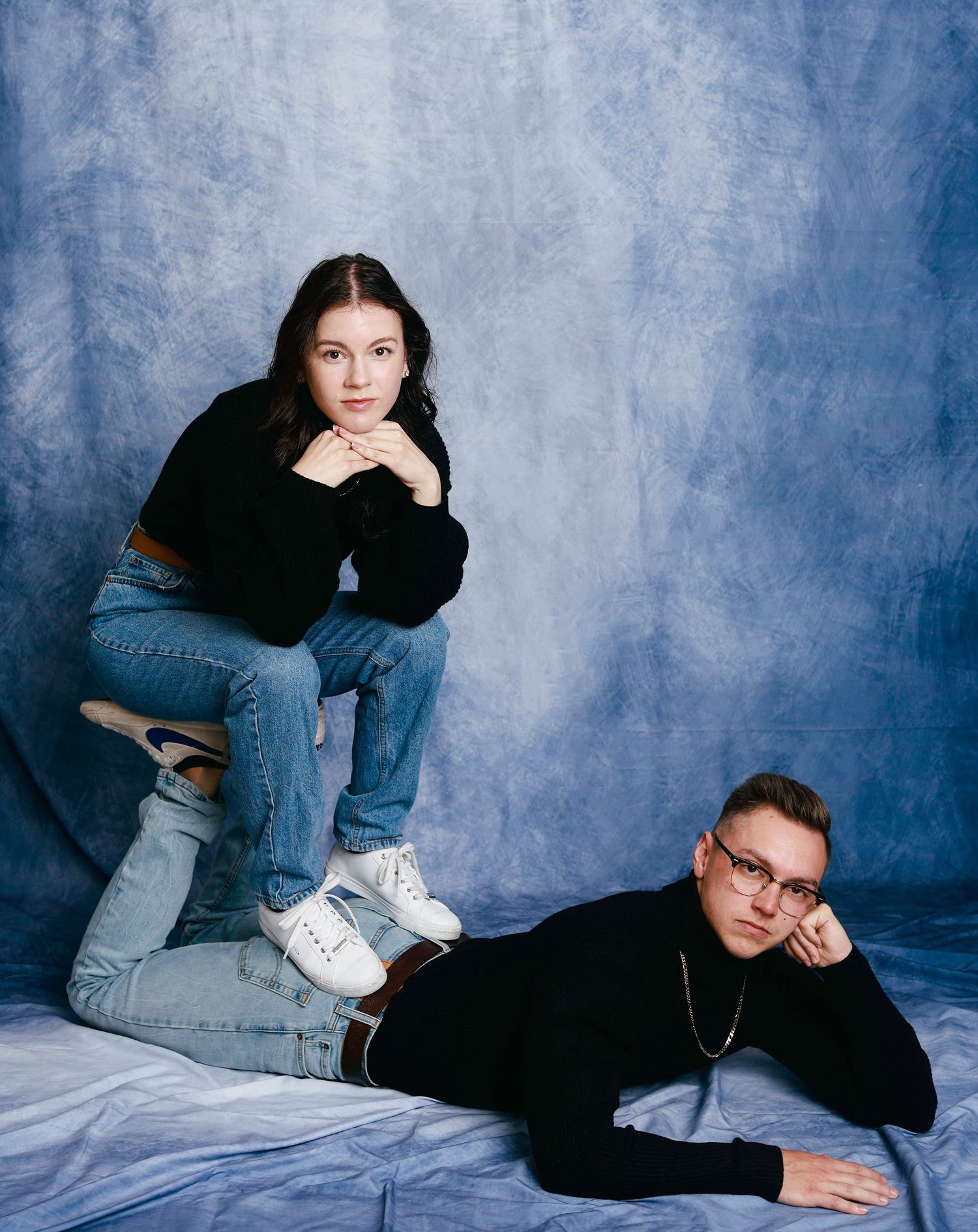 Portrait of two people in black sweaters and jeans posed against a mottled blue backdrop in a casual studio setting.