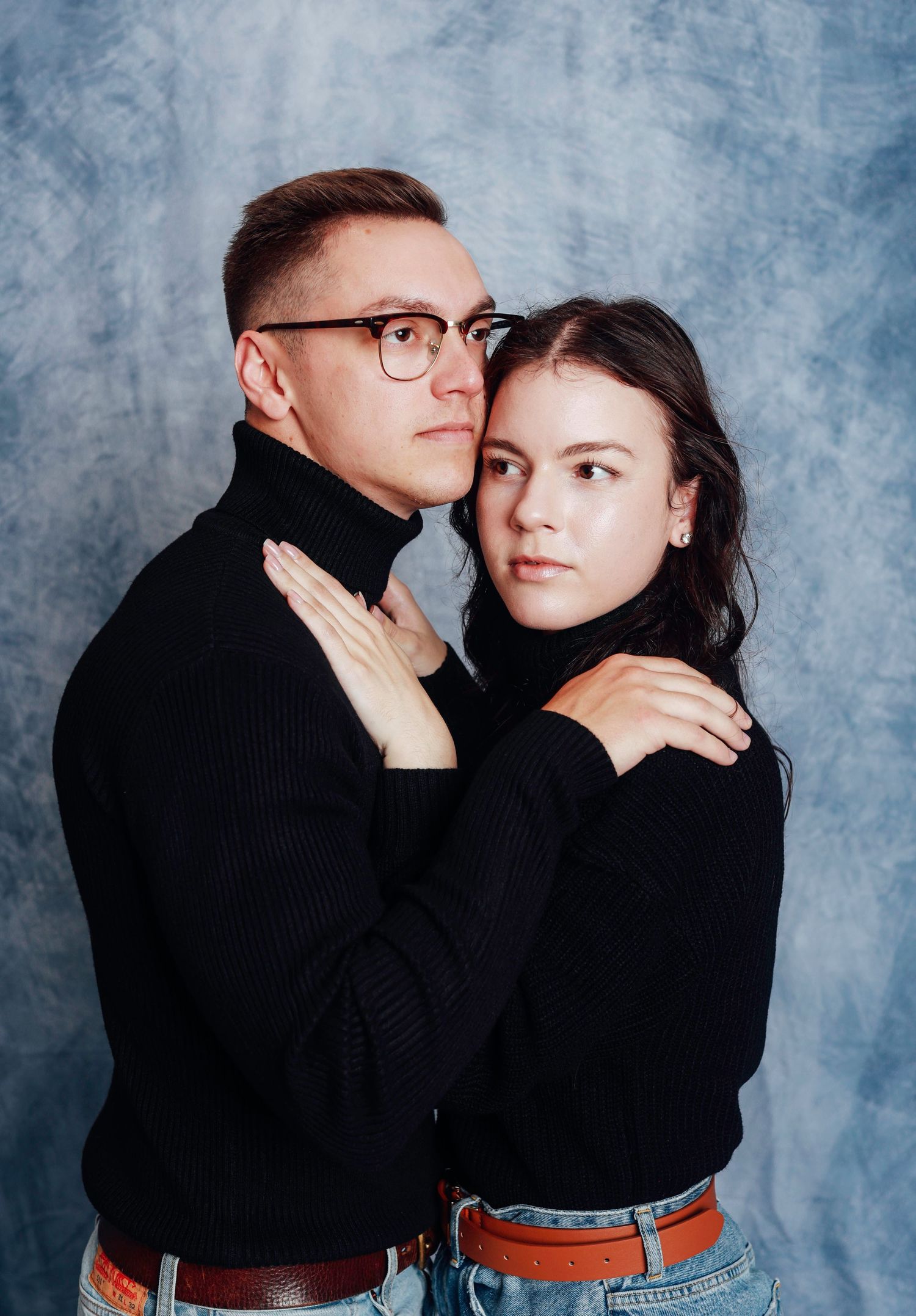 Romantic studio portrait of a couple in matching black sweaters posing against a textured blue background.