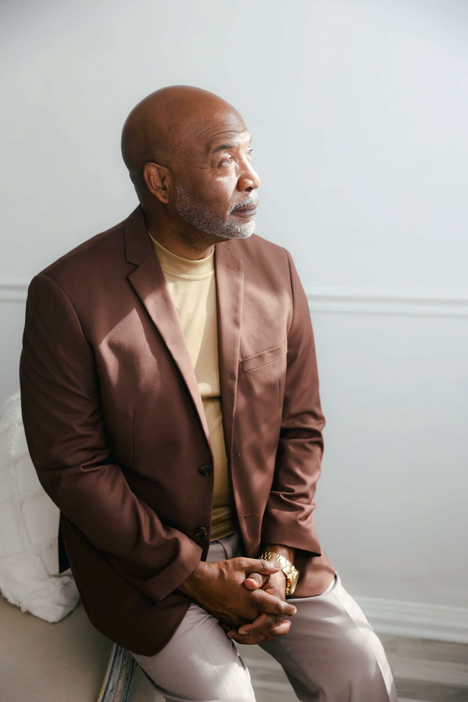 A person wearing a brown blazer and beige shirt sits thoughtfully in a minimalist white setting.