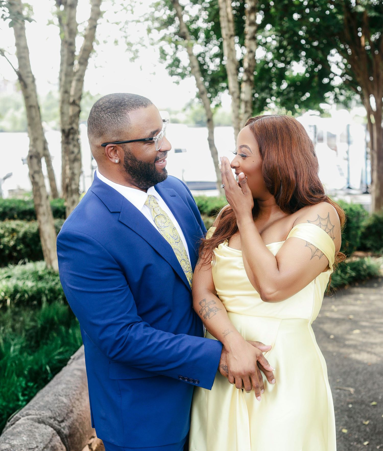 A couple in yellow and blue formal attire laugh and embrace while enjoying a romantic moment outdoors among trees.