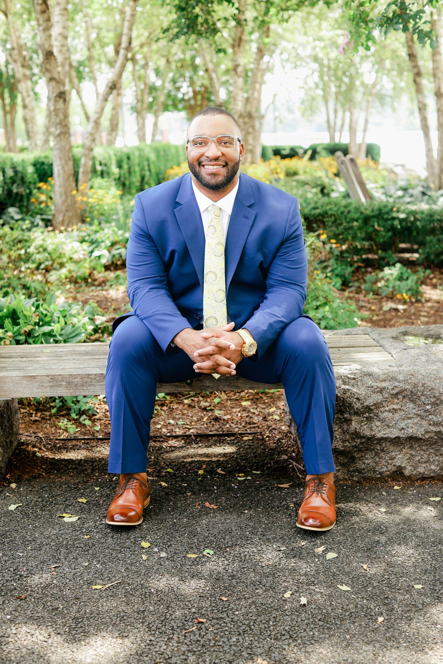 Professional wearing a navy suit sits on stone wall surrounded by lush greenery, showing polished brown leather shoes.