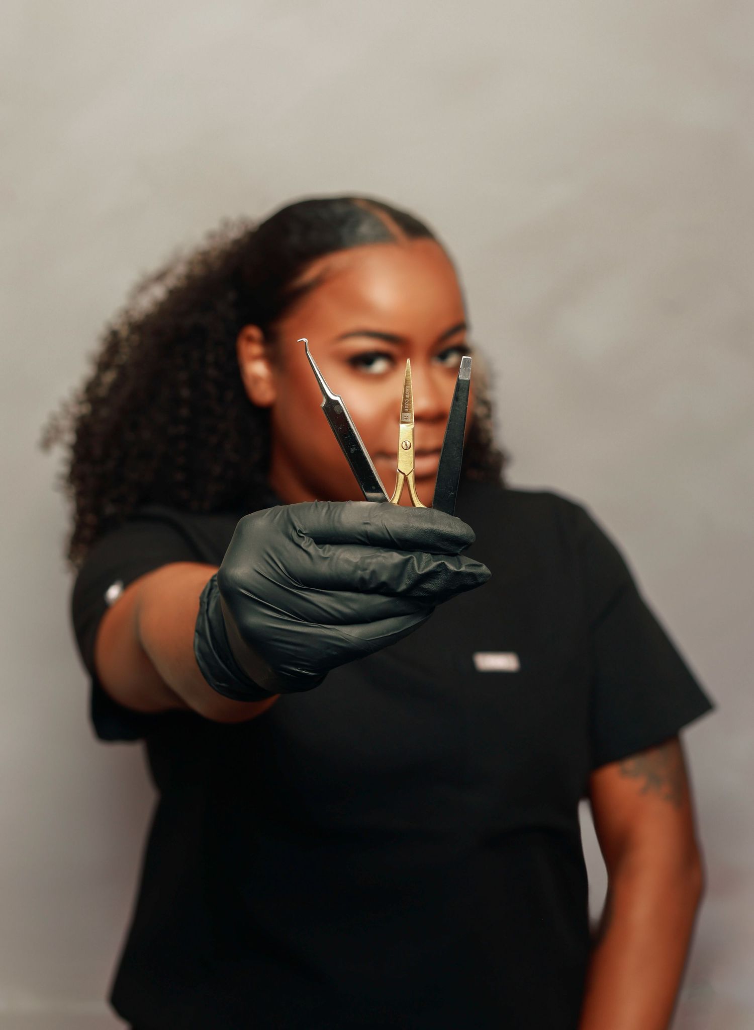 Beauty professional wearing black gloves holds gold measuring tool against neutral background.
