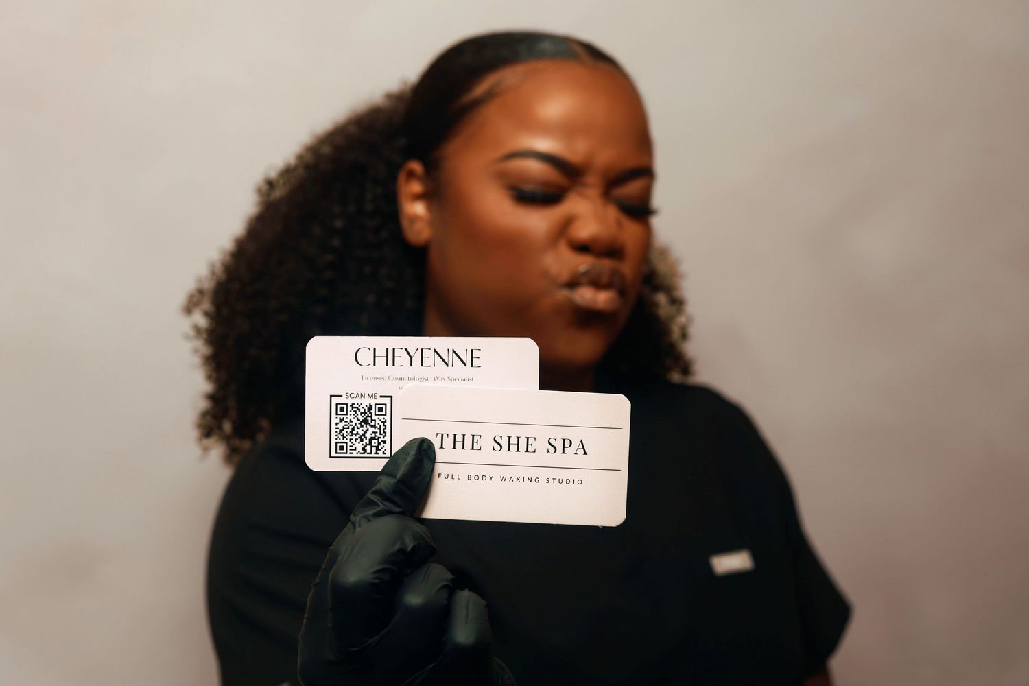 A person wearing black gloves holds a The She Spa business card from Cheyani against a neutral background.