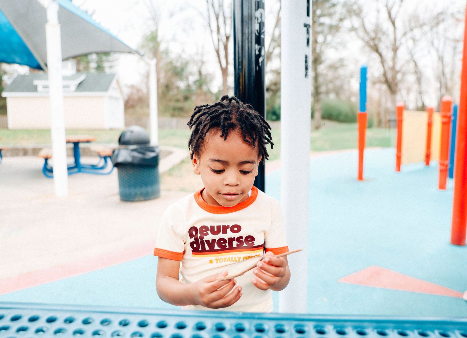 A child wearing a striped shirt uses a mobile device while sitting at a playground with colorful equipment in the background.
